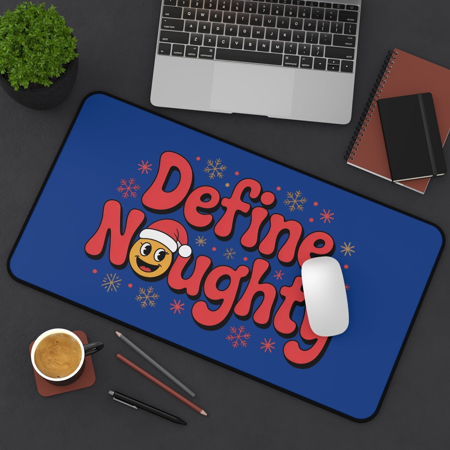 Define Naughty Desk Mat with vibrant blue background, playful red lettering, and Santa emoji, placed on a black desk with laptop, coffee, and stationery.