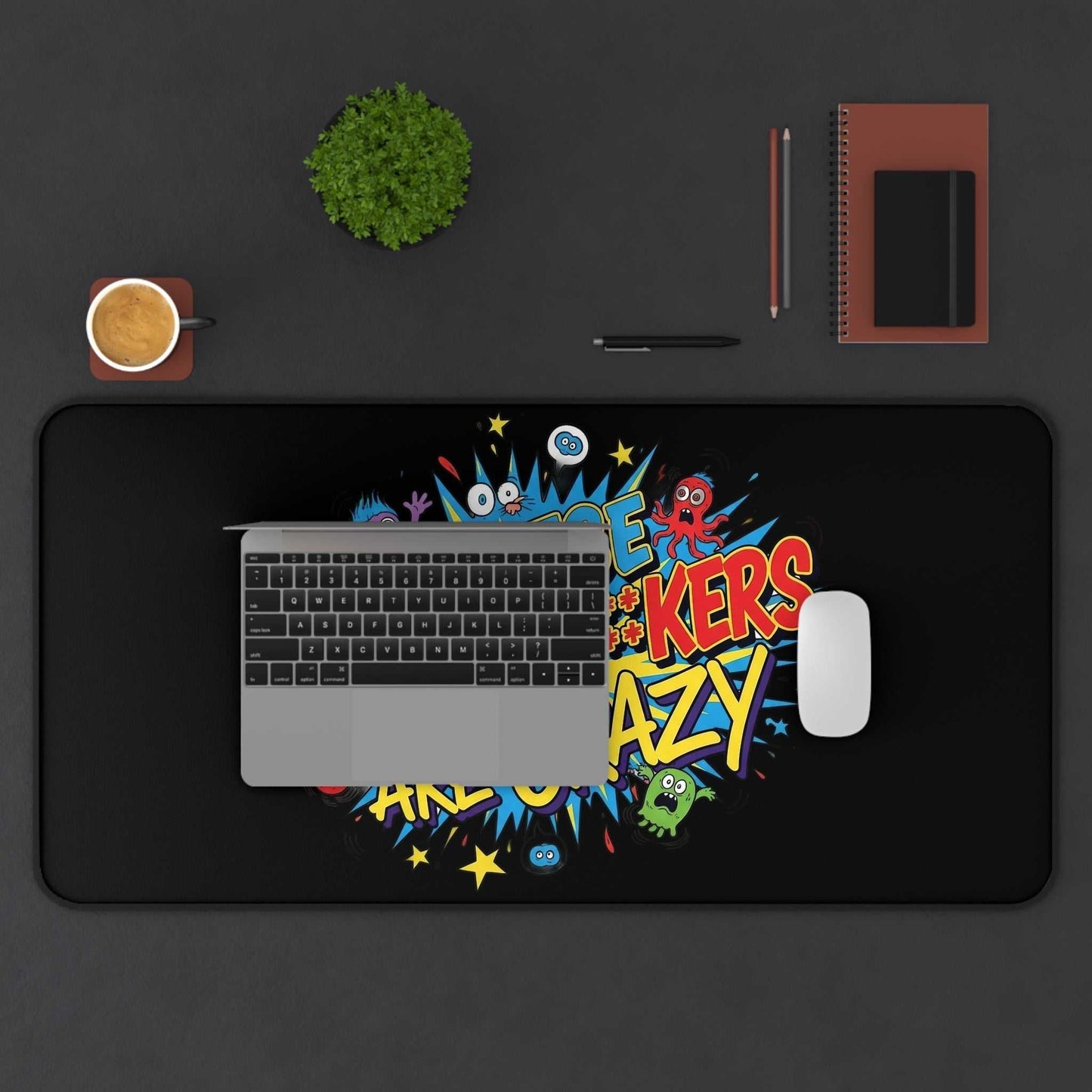Bold colorful desk pad with humorous "These Motherf++kers Are Crazy" text, non-slip bottom, and hemmed edges on a workspace with laptop, mouse, coffee, and stationery.