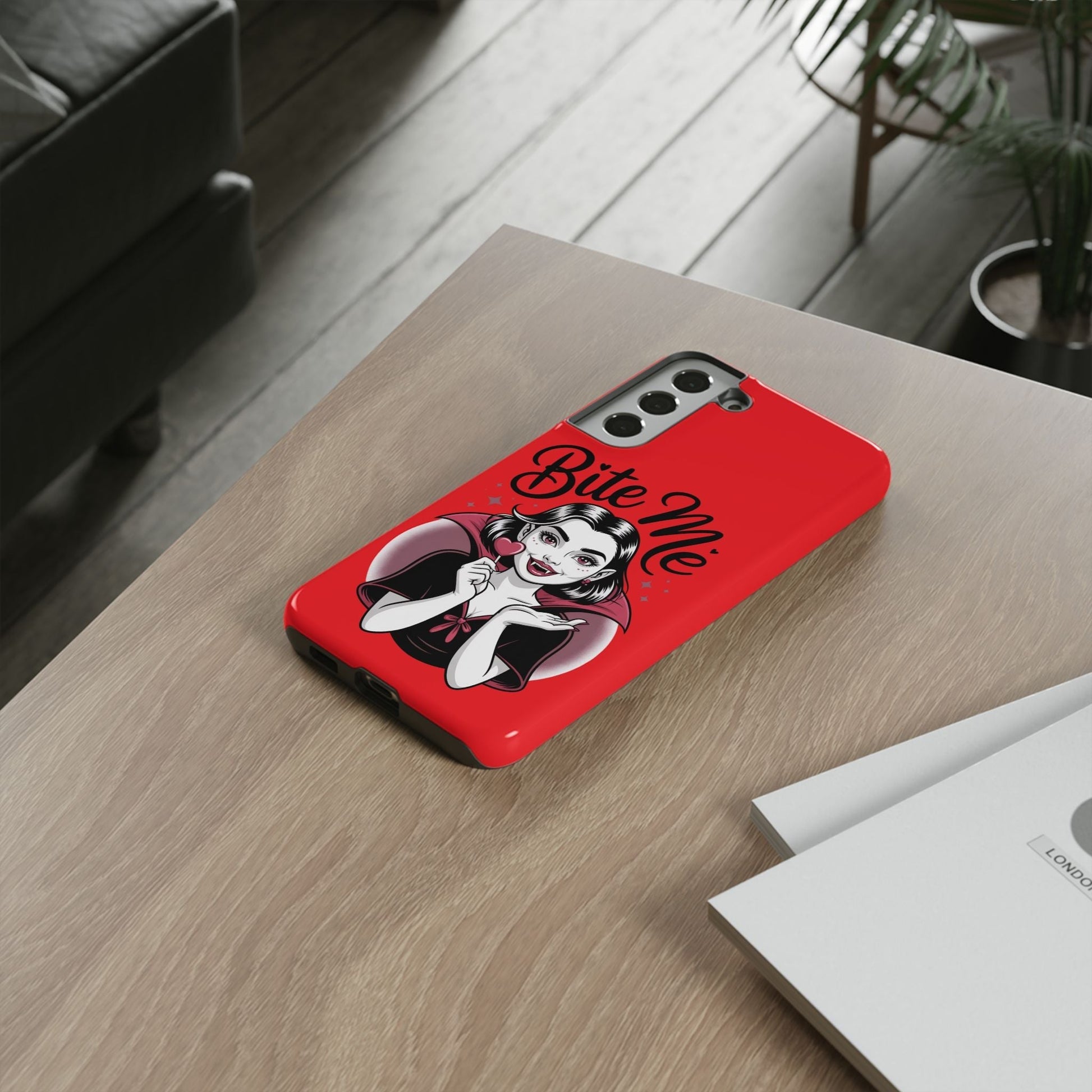 Bright red Bite Me Vampire Samsung phone case featuring a cartoon vampire design and bold text on a wooden table