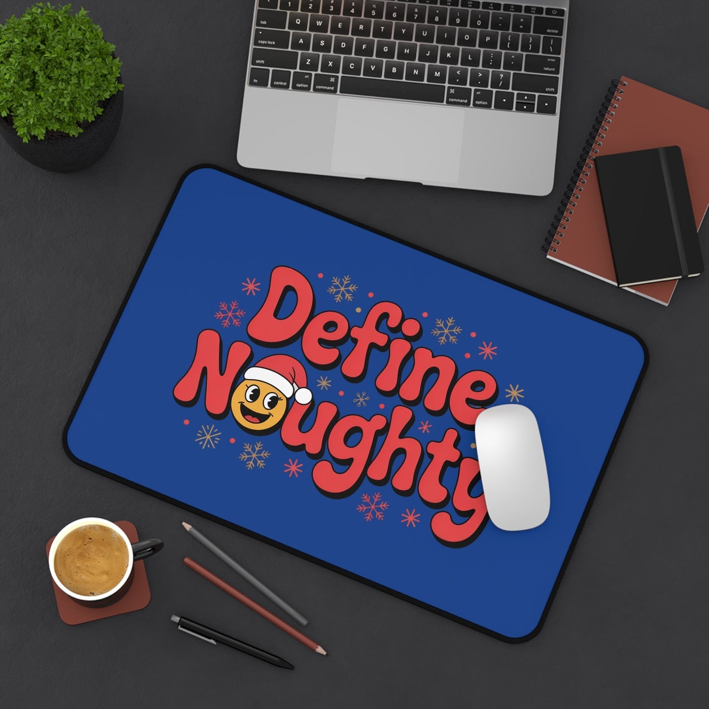 Define Naughty Desk Mat with playful holiday design on blue background, featuring red text and emoji with Santa hat, placed on workspace with laptop, mouse, coffee, and notebooks