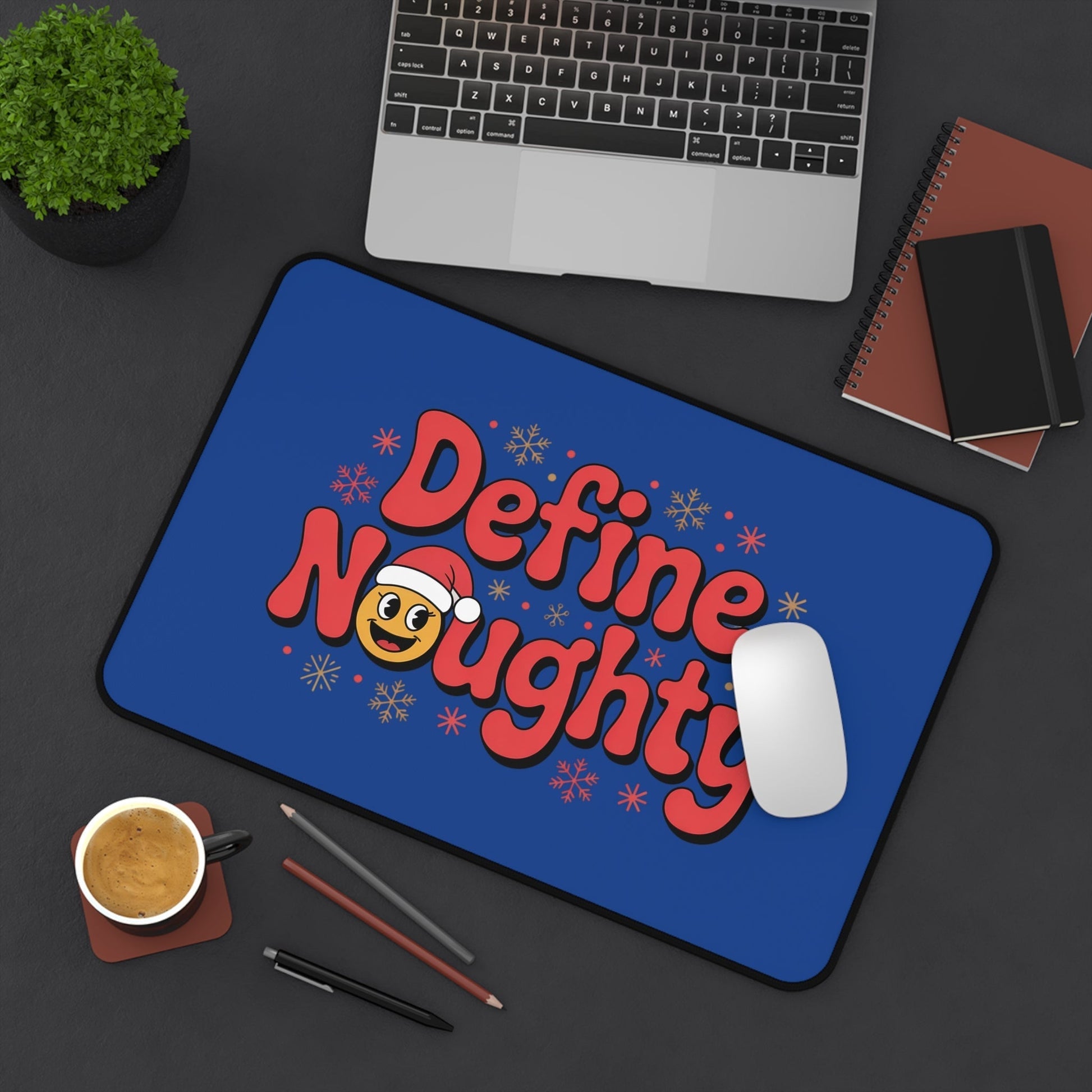 Define Naughty Desk Mat with playful holiday design on blue background, featuring red text and emoji with Santa hat, placed on workspace with laptop, mouse, coffee, and notebooks